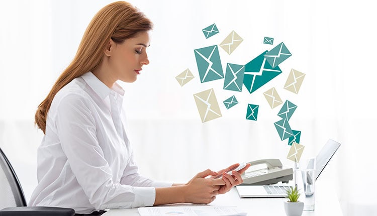 A woman sits at a desk using her phone, with illustrated envelopes appearing to flow from her device toward a laptop, symbolizing email communication.