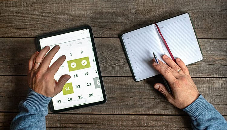 Person with a blue sweater schedules events using a digital calendar on a tablet and writes in an open paper planner on a wooden table.