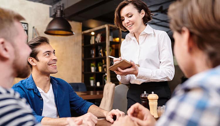 A waitress takes an order from three seated customers at a restaurant, smiling and holding a notepad.