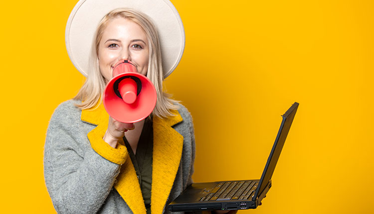 A woman wearing a hat and coat holds a laptop in one hand and speaks into a red megaphone against a yellow background.