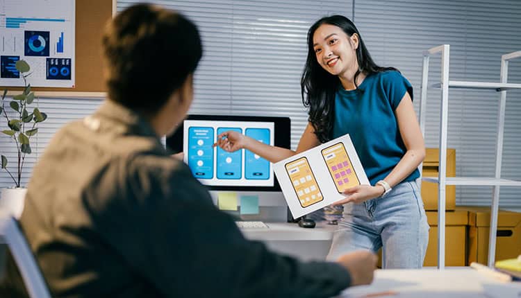 Woman presenting app interface designs on paper to a seated colleague in an office with screens displaying similar designs in the background.