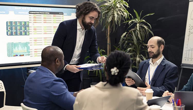 Four business professionals sit around a meeting table while one man hands out documents; a large screen showing charts and data is visible in the background.