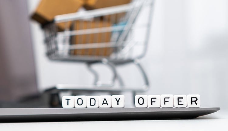 Close-up of letter blocks spelling "TODAY OFFER" on a laptop, with a miniature shopping cart and packages in the background.