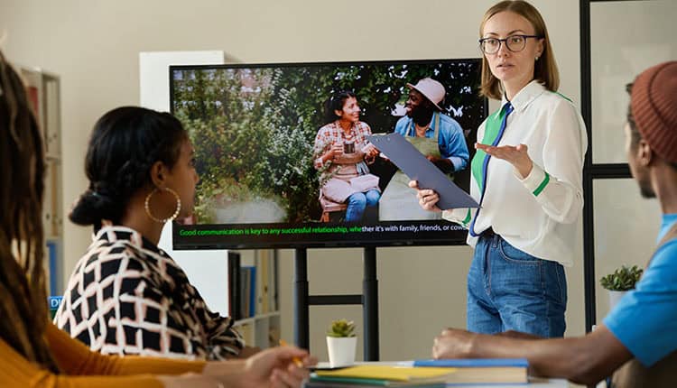 A woman stands in front of a screen presenting to three seated people; the screen displays an image of two people talking outdoors with a caption about communication.
