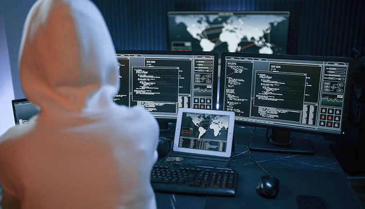 Person in a hoodie sits at a desk with multiple monitors displaying code and a world map